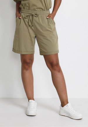 Persoon draagt olijfgroene shorts met trekkoord en zakken, samen met witte sneakers, staand tegen een witte achtergrond.