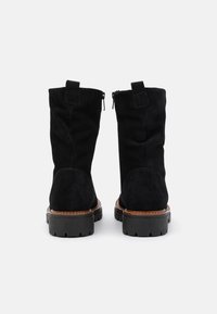 Botas de tobillo de ante negro con una suela de goma negra gruesa, que presentan cremalleras laterales y un tacón marrón cosido de forma contrastante.