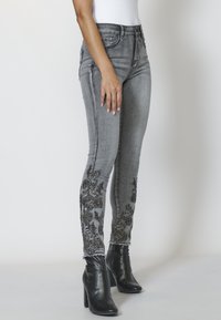 Šedé skinny džíny z denimu s květinovou výšivkou na kotnících, roztrhaným lemem a přiléhavým střihem. Doplněno černými kotníkovými botami.