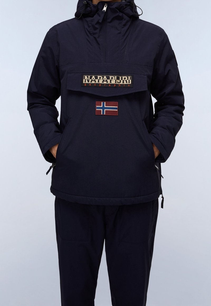 Personne portant une veste Napapijri bleu marine avec une grande poche avant, un écusson du drapeau norvégien, les mains dans les poches latérales, et un pantalon assorti bleu marine.