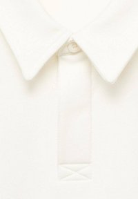 Valkoinen polo paita, jossa on pehmeä pinta, taitettu kaulus ja kaksinappinen kiinnitys. Kauluksen alla on hienovarainen kolmioommel yksityiskohta.