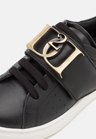 Sneaker in pelle nera con una grande fibbia oro, lacci neri e dettagli testurizzati ai lati, caratterizzato da una suola bianca.