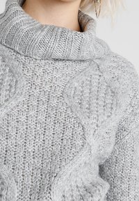 Grauer Strickpullover mit hohem Kragen, strukturierten Mustern und Zopfmuster-Details. Das Material wirkt weich und gemütlich.