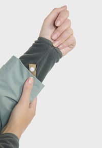 Manche en polaire de couleur vert foncé s'étendant à partir d'une poche imperméable vert clair, avec un onglet beige et un fermoir à pression blanc.