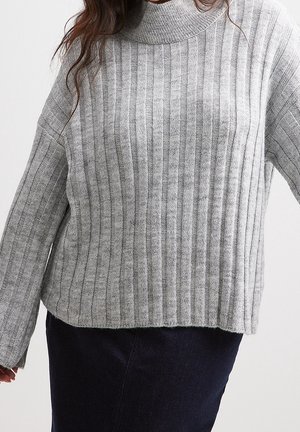 Kobieta ubrana w jasnoszary prążkowany sweter z golfem i długimi rękawami, zestawiony z ciemnymi spodniami.