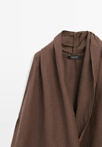 Vêtement marron en lin à manches longues avec col châle suspendu, montrant l’étiquette "Massimo Dutti Made in Morocco" à l’intérieur du col.