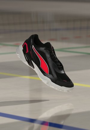 Schwarzer Sportschuh mit rotem Design und weißer Sohle, der über einem Hallenboden mit farbigen Linien schwebt.