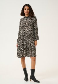 Zwarte en crèmekleurige jurk met een patroon, lange mouwen, een gerimpelde taille, een gelaagd rok en een hoge halslijn, gecombineerd met zwarte enkelboots.
