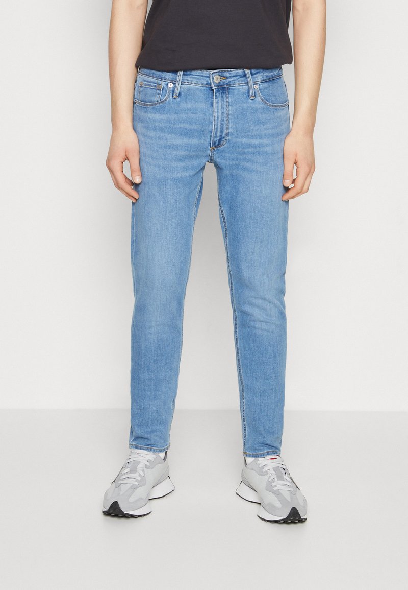 Jeans de mezclilla azul claro con un corte slim. Presentan un diseño clásico de cinco bolsillos, un ligero desgaste y costuras en contraste. Llevados con zapatillas deportivas grises.