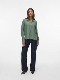 Blouse vert clair à manches longues en tissu lisse avec un devant à boutons, associée à un jean taille haute bleu foncé à jambes larges et des escarpins pointus noirs à pois.