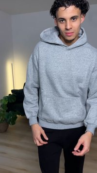 Sweat à capuche gris en tissu doux, avec une poche avant et un cordon de serrage à la capuche. Porté avec un pantalon noir dans un environnement intérieur bien éclairé.