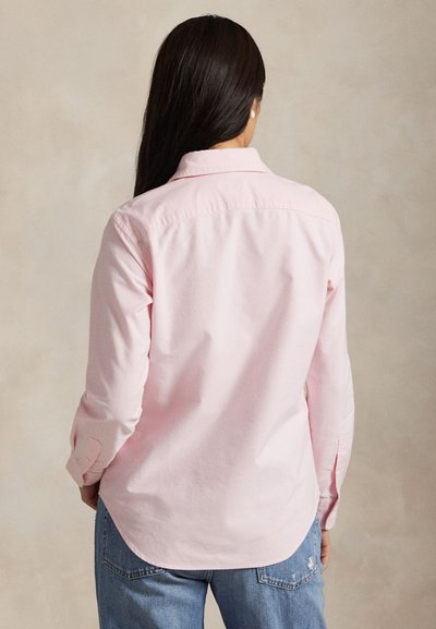 Frau mit langen dunklen Haaren trägt ein hellrosa langärmeliges Shirt und blaue Jeans, von hinten vor einem einfarbigen Hintergrund gezeigt.
