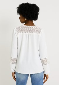 Blusa blanca de manga larga con intrincados detalles de encaje en los hombros y los puños; tejido texturizado y un ajuste relajado.