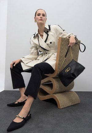 Gabardina beige con ribetes negros, pantalones negros cropped, zapatos negros de tiras con detalle dorado y un bolso negro texturizado sobre una silla curvada.