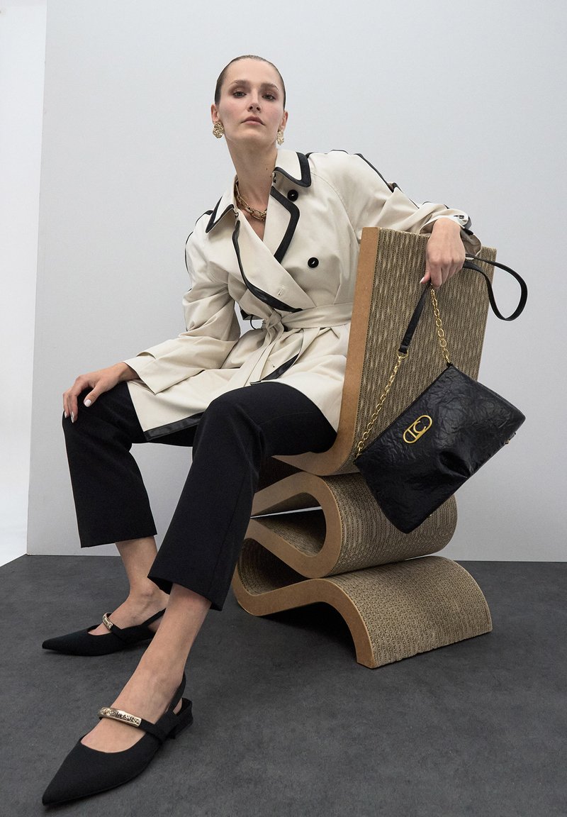Gabardina beige con ribetes negros, pantalones negros cropped, zapatos negros de tiras con detalle dorado y un bolso negro texturizado sobre una silla curvada.