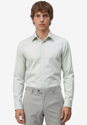 Junger Mann trägt ein hellgrünes, körperbetontes Hemd, das in hellgraue, maßgeschneiderte Hosen gesteckt ist, und steht vor einem schlichten Hintergrund.