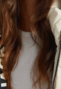 Hvid ribbet top under en dunjakke med lynlås og sorte detaljer; bølget, langt brunt hår rammer halsudskæringen.