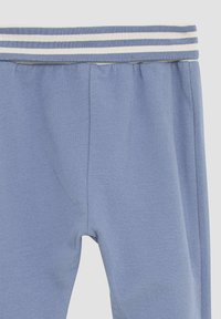 Jasnoniebieskie dresy o miękkiej teksturze, z prążkowanym pasem w talii z białymi paskami i luźnym krojem. Brak widocznych kieszeni.