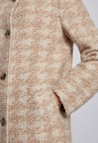 Beige og cremefarvet tekstureret uldfrakke med knapper, med en frontlomme hvor en hånd delvist er indsat.