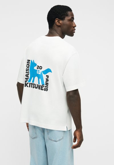 Άνδρας που φορεί λευκό υπερμέγεθες μπλουζάκι με μπλε γραφικό αλεπούς και κείμενο "Maison Kitsuné Paris 2002" στην πλάτη, συνδυασμένο με ανοιχτό μπλε τζιν.