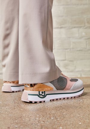 Gros plan d’une personne portant des baskets rose clair et grises avec des accents en or rose et un pantalon beige, debout sur un sol texturé.