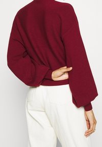 Mörkröd stickad tröja med lös passform, sänkta axlar och ribbade ärmslut. Kombinerad med högmidjade vita byxor. Texturerad tygdetalj synlig.