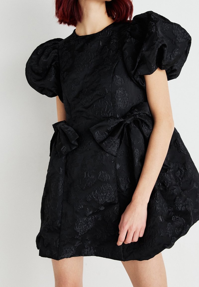 Femme portant une robe noire texturée avec des manches bouffantes, un motif floral et deux grands nœuds sur le devant, debout devant un fond uni.