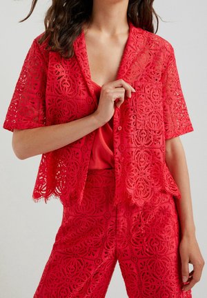 Frau trägt rotes Spitzen-Kurzarmshirt und passende Hose mit floralen geometrischen Mustern, hält teilweise den Hemdkragen.