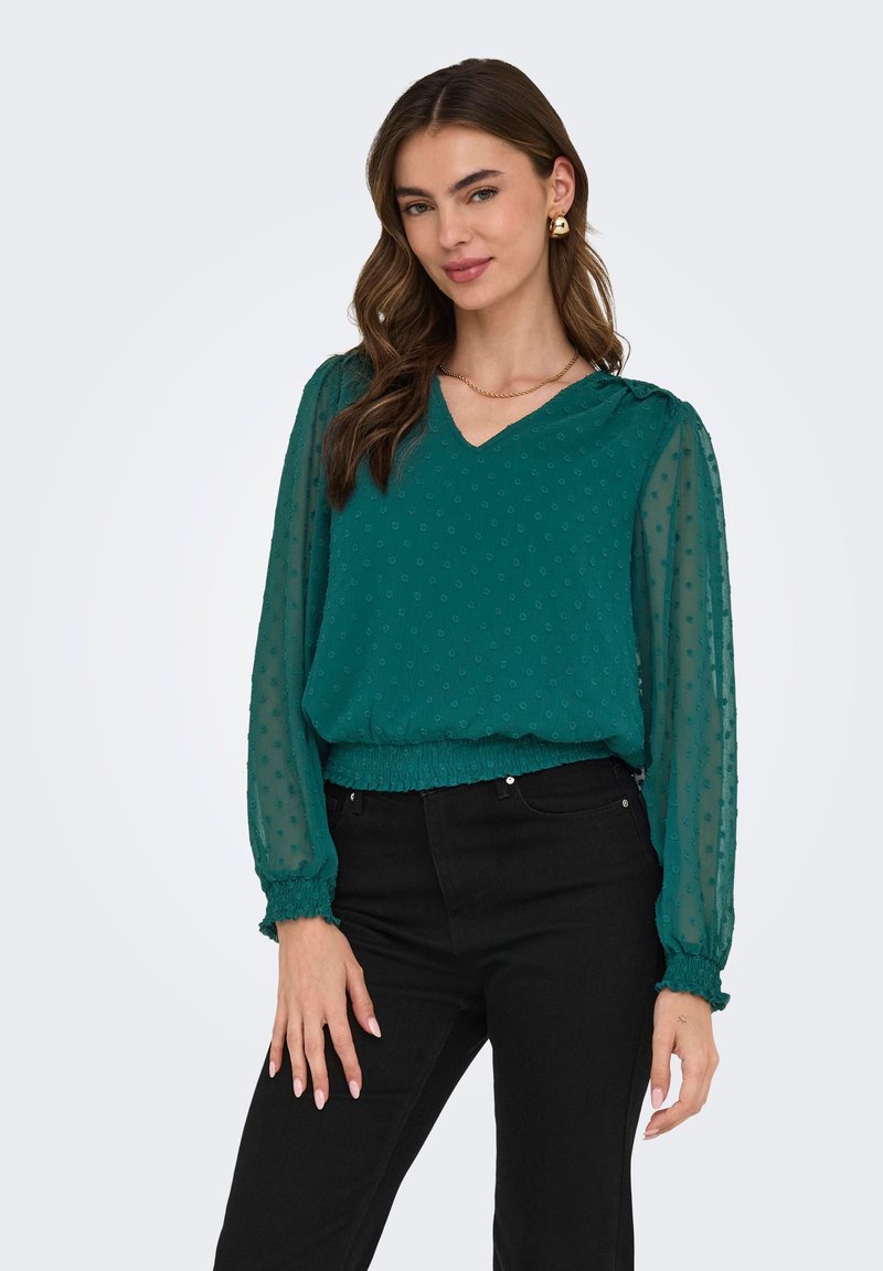 Femme portant un chemisier à manches longues texturé couleur sarcelle avec taille et poignets froncés, associé à un pantalon noir, posant devant un fond blanc.