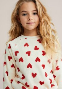 Bluză de fleece cu bază crem și modele cu inimi roșii, având guler rotund și mâneci raglan, textură moale și croială lejeră.