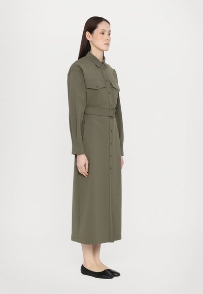 Olivegrünes Longsleeve-Hemdkleid mit Knopfverschluss, zwei Brusttaschen, tailliertem Gürtel und gerader Silhouette; kombiniert mit schwarzen Ballerinas.