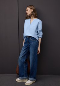 Chemise bleu clair à manches longues avec un col en V et des poignets retroussés, associée à un jean en denim bleu à jambes larges et des baskets blanches. Sac marron visible.