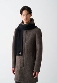 Brun uldfrakke med høj krave, parret med et langt, ribbet sort halstørklæde. Halstørklædet har et rundt logo i enden.