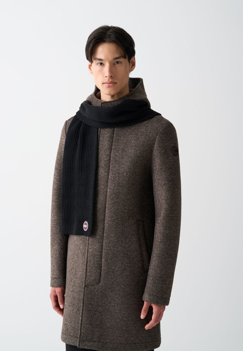 Brun uldfrakke med høj krave, parret med et langt, ribbet sort halstørklæde. Halstørklædet har et rundt logo i enden.