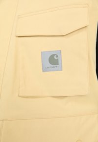Geltona drobės darbo liemenė su dideliu krūtinės kišeniu ir kvadratiniu Carhartt logotipu pilkos spalvos. Lygus paviršius su sustiprintomis siūlėmis.