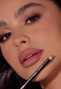 Matowy ołówek do ust trzymany blisko pełnych warg w delikatnym różowym odcieniu. Skóra jest gładka, podkreślona wokół oczu, z wyrazistymi rzęsami i zdefiniowanymi brwiami.