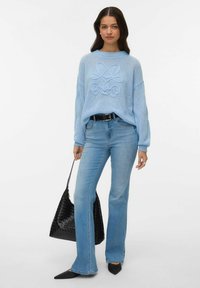 Svetlo modra pletena majica z teksturiranim vzorcem, združena s svetlo modrimi razširjenimi jeans hlačami, črno tkanim torbico in črnimi špičastimi balerinkami.