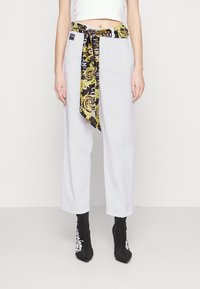 Pantalones recortados blancos de cintura alta con grandes bolsillos frontales, combinados con botas de tobillo negras que presentan texto blanco y un cinturón de bufanda negro y amarillo con un patrón.