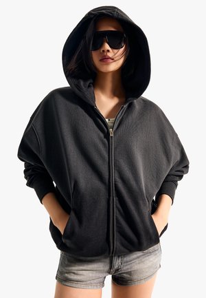 Frau trägt übergroßen schwarzen Hoodie mit Reißverschluss und aufgesetzter Kapuze, schwarze Sonnenbrille und helle Jeansshorts, Hände in den Taschen vor weißem Hintergrund.