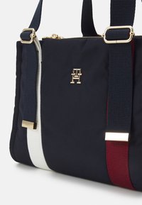 Granatowa torba wykonana z materiału typu tote z zapięciami w kolorze złotym, z widocznym logo. Elementy dekoracyjne to białe i bordowe paski po bokach oraz teksturowane paski.