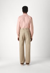 Ljusrosa bomullsskjorta med lång ärm, kombinerad med beige strukturerade vida byxor och bruna skor. Utsikt bakifrån, enkel bakgrund.