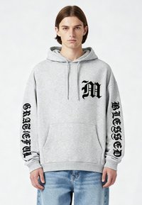 Felpa grigia in pile con tasca a marsupio. Presenta la stampa nera della scritta "M" e "GRATEFUL" sulle maniche e "BLESSED" sul petto.