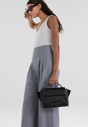 Black leather handbag with a structured design, top handle and zippered sides. Model wears gray pleated trousers and a white tank top.