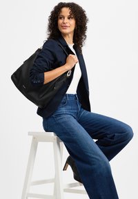 Schwarze Handtasche aus genarbtem Leder mit goldfarbenen Beschlägen, ausgestattet mit einem Reißverschluss und seitlichen Taschen, kombiniert mit einem marineblauen Blazer und Jeans.