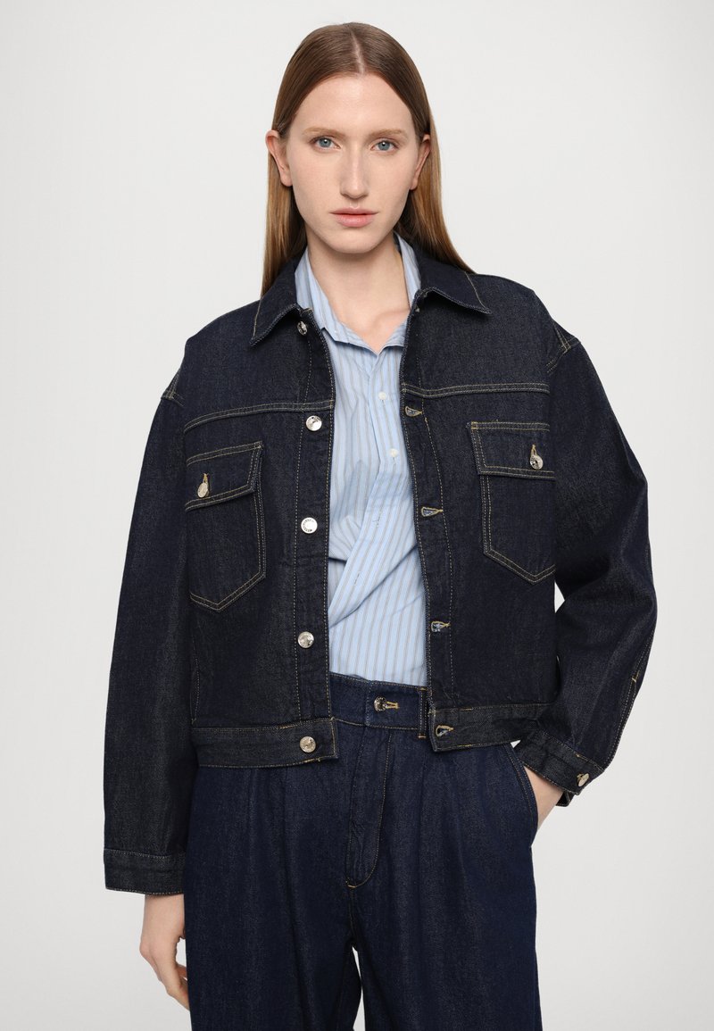 Giacca in denim scuro con design accorciato, bottoni a pressione argentati, due tasche sul petto e cuciture a contrasto. Indossata sopra una camicia a righe blu chiaro.