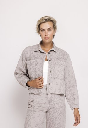 Blonde Frau trägt eine hellgraue Jeansjacke mit Leopardenmuster und passende Hose und posiert mit einer Hand, die die Vorderseite der Jacke hält.