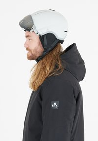 Hvid skihjelm med mørkt visir, sort fleece-for og justerbar hagerem. Båret med en sort jakke med et logo-patch på ærmet.