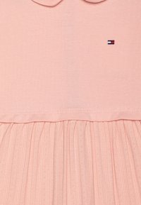 Ljusrosa klänning med en texturerad överdel med krage och en plisserad kjol. Litet logotyp i röd, vit och blå på bröstet.