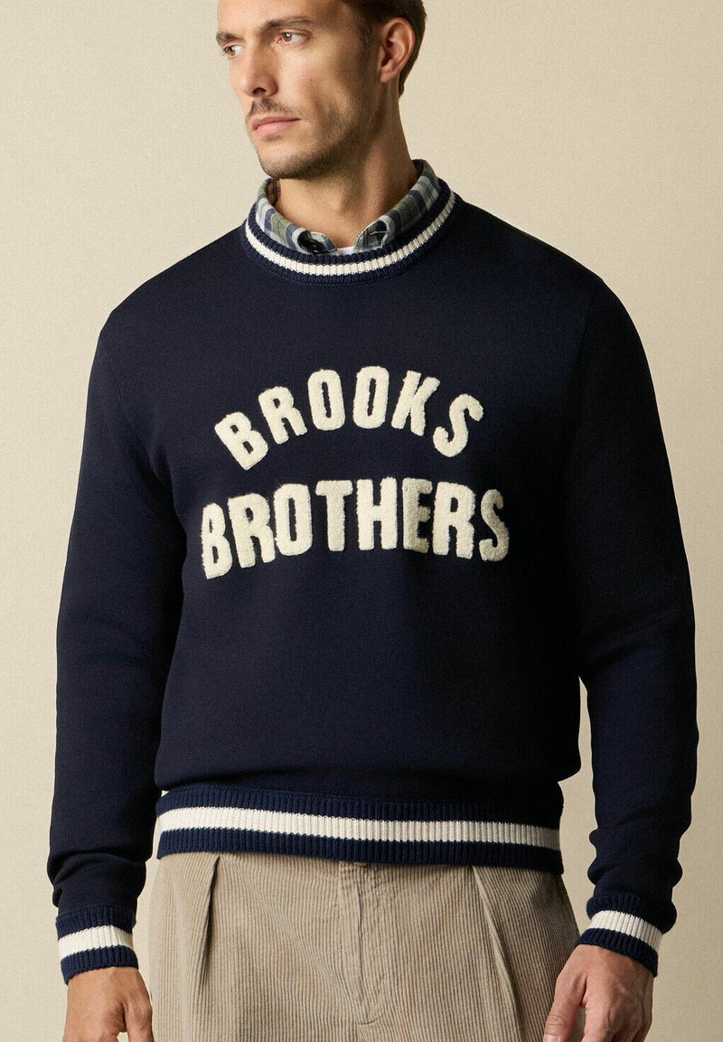 Námořnický vlněný svetr s nápisem "BROOKS BROTHERS" v krémové barvě. Má žebrované manžety a lem s bílými a námořnickými pruhy.
