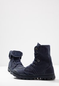 Botas de tobillo de lona azul marino con suelas de goma, diseño de cordones y una solapa plegable en el cuello. Presenta ojales de metal y tela texturizada.